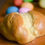 A rustic loaf of sweet Paska bread sits on a cutting board with colourful Easter eggs in the background.