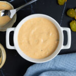 Burger sauce in a white bowl, next to a jar, and spoon on a lid, pickles, and a blue napkin.