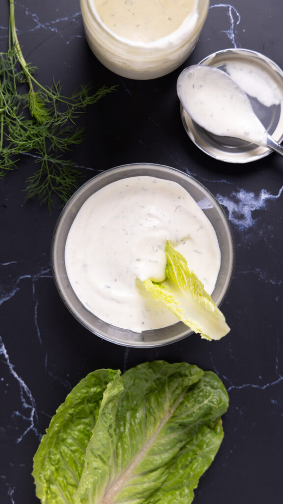 Overhead photo of a bowl of ranch dressing, next to an open jar, a spoon on a lid, a sprig of fresh drill, and leaves of romaine lettuce.