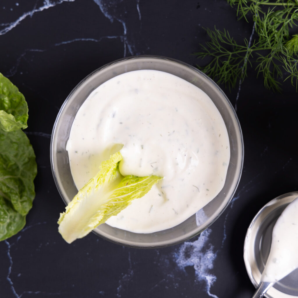 Overhead photo of a bowl of ranch dressing, next to an open jar, a spoon on a lid, a sprig of fresh drill, and leaves of romaine lettuce.