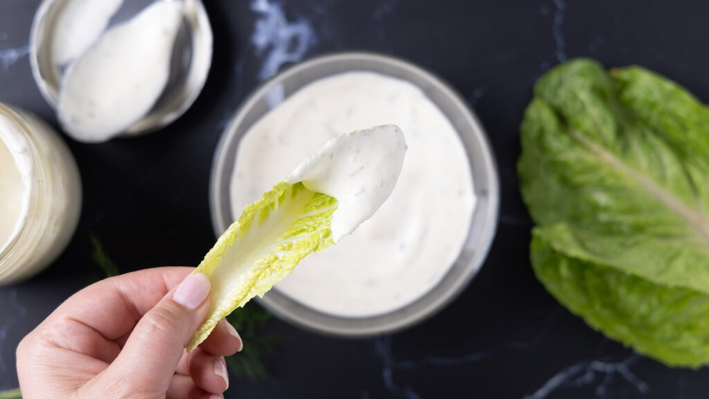 Overhead photo of a bowl of ranch dressing, next to an open jar, a spoon on a lid, a sprig of fresh drill, and leaves of romaine lettuce.