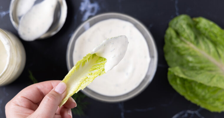 Overhead photo of a bowl of ranch dressing, next to an open jar, a spoon on a lid, a sprig of fresh drill, and leaves of romaine lettuce.