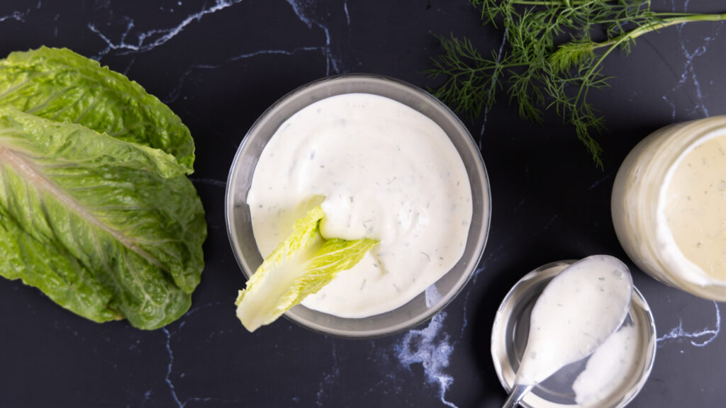 Overhead photo of a bowl of ranch dressing, next to an open jar, a spoon on a lid, a sprig of fresh drill, and leaves of romaine lettuce.