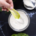 Overhead photo of a bowl of ranch dressing, next to an open jar, a spoon on a lid, a sprig of fresh drill, and leaves of romaine lettuce.