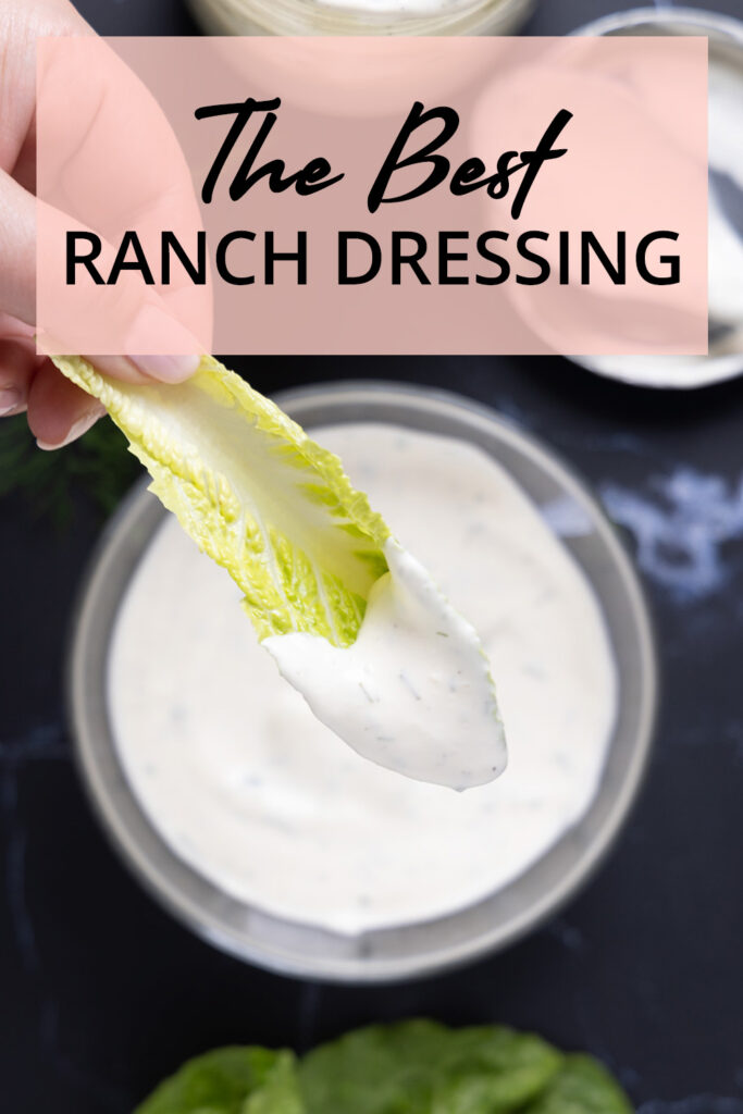 Overhead photo of a bowl of ranch dressing, next to an open jar, a spoon on a lid, a sprig of fresh drill, and leaves of romaine lettuce.