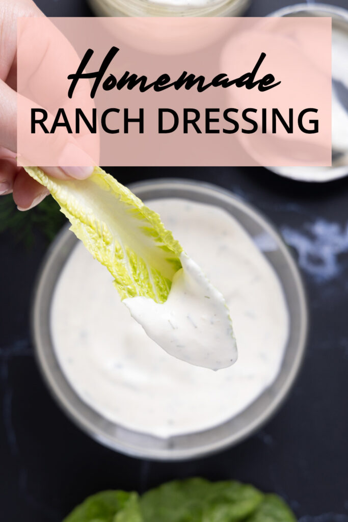 Overhead photo of a bowl of ranch dressing, next to an open jar, a spoon on a lid, a sprig of fresh drill, and leaves of romaine lettuce.