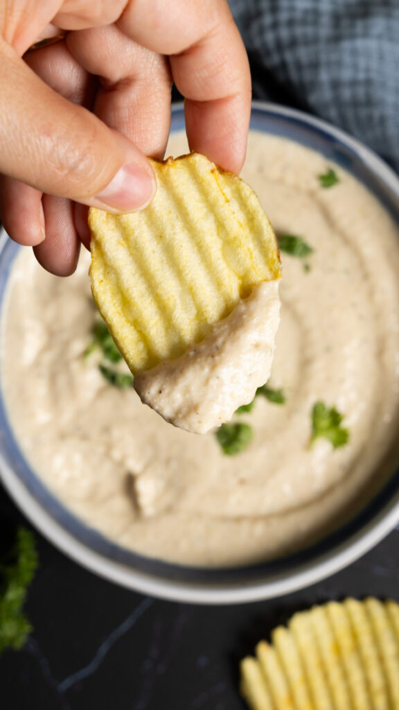 A bowl of onion dip sits on a counter with ripple potato chips on the side. A hand holding a ripple potato chip has scooped some dip onto the chip.