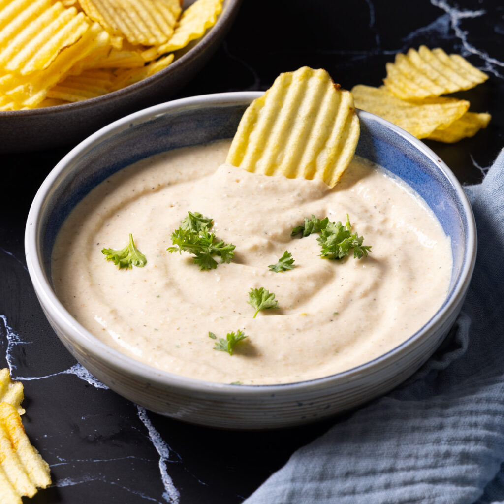 A bowl of onion dip sits on a counter with ripple potato chips on the side. 