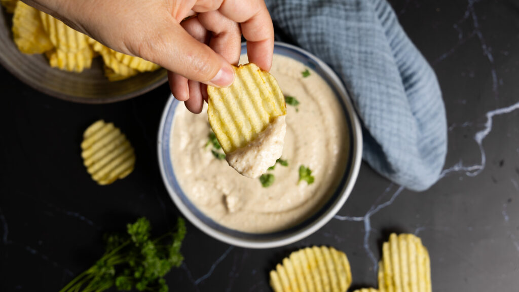 A bowl of onion dip sits on a counter with ripple potato chips on the side. A hand holding a ripple potato chip has scooped some dip onto the chip.