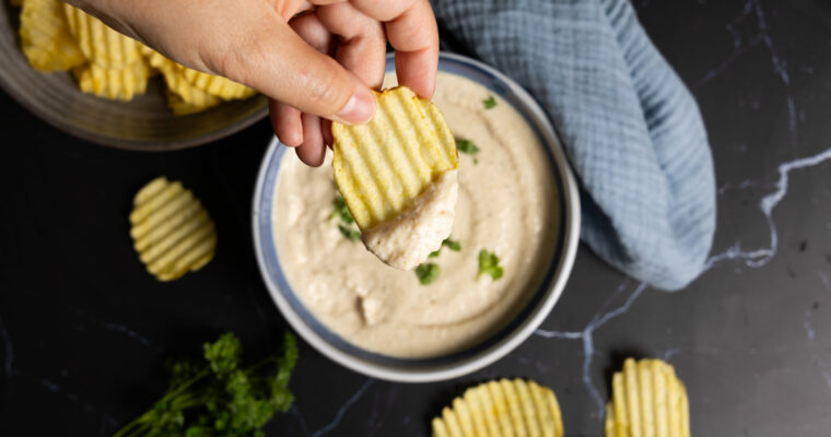 A bowl of onion dip sits on a counter with ripple potato chips on the side. A hand holding a ripple potato chip has scooped some dip onto the chip.