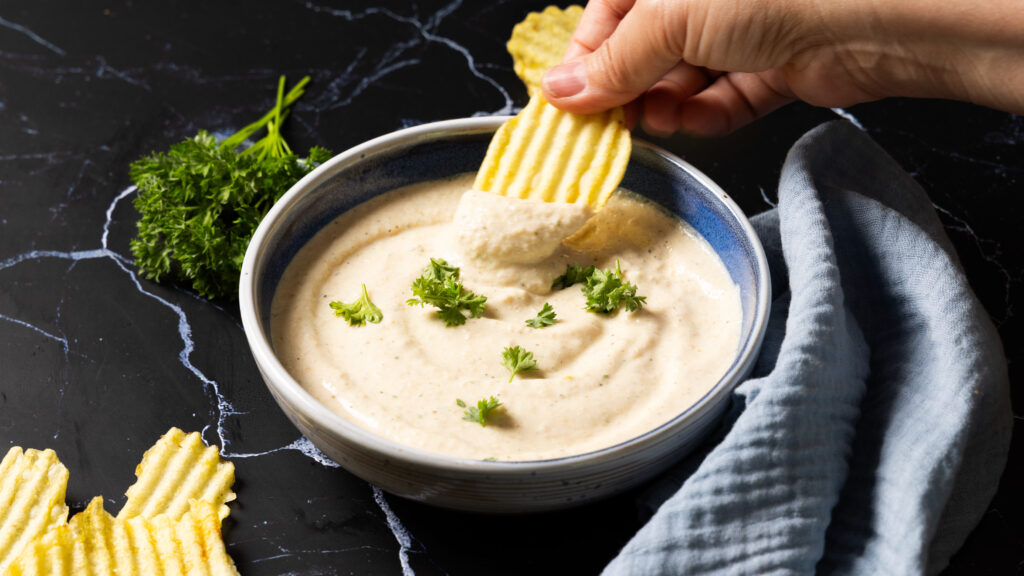 A bowl of onion dip sits on a counter with ripple potato chips on the side. A hand holding a ripple potato chip has scooped some dip onto the chip.