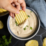 A bowl of onion dip sits on a counter with ripple potato chips on the side. A hand holding a ripple potato chip has scooped some dip onto the chip.