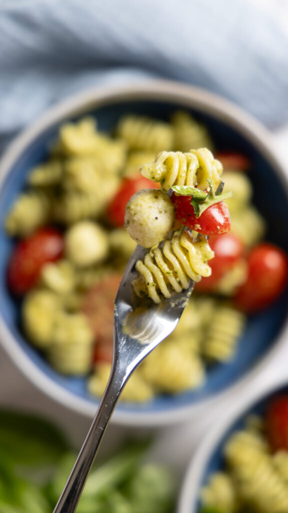 A pasta salad in a large salad bowl, with small tomatoes, basil pesto sauce, mini mozzarella balls, and chopped fresh basil. A fork is lifting a bite of salad.