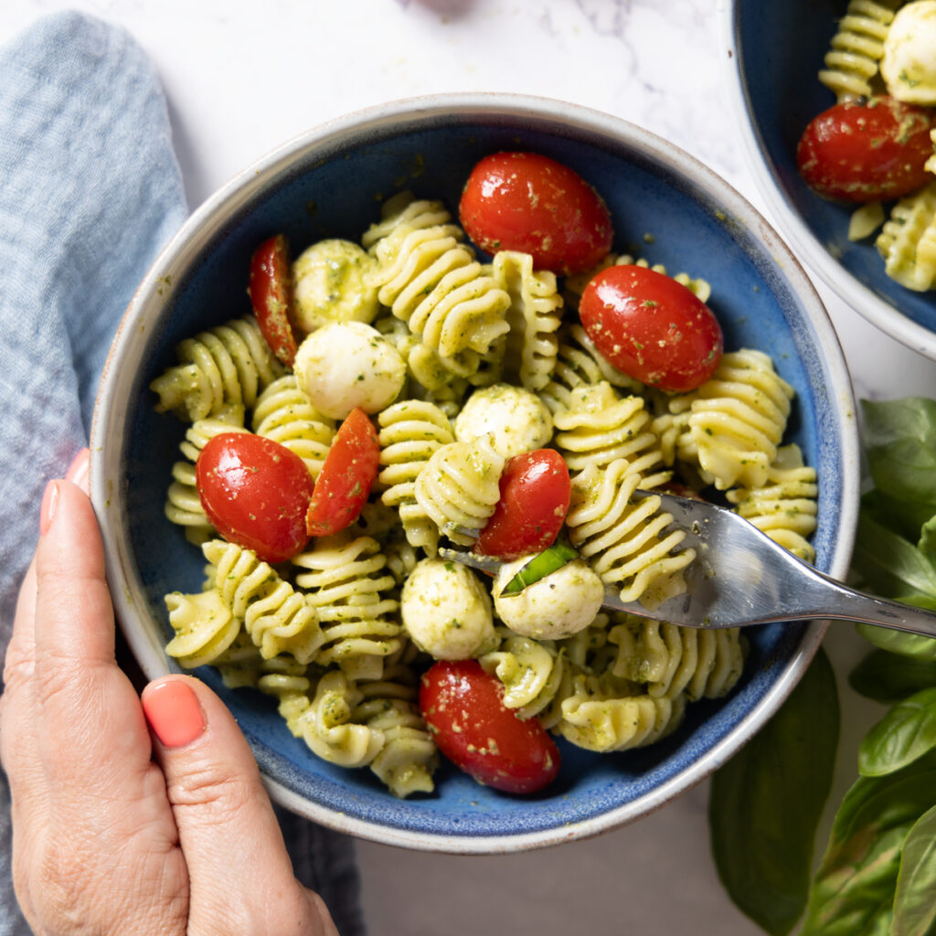 A pasta salad in a large salad bowl, with small tomatoes, basil pesto sauce, mini mozzarella balls, and chopped fresh basil. Hands are holding the bowl and a fork is lifting a bite of salad.