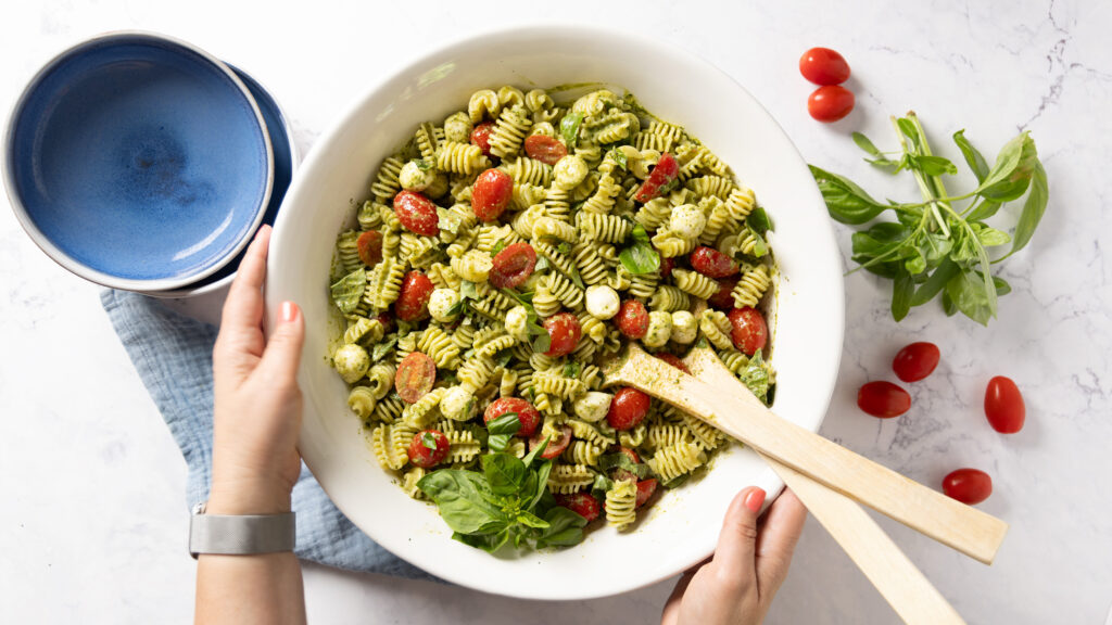 A pasta salad in a large salad bowl, with small tomatoes, basil pesto sauce, mini mozzarella balls, and chopped fresh basil. Hands are holding the bowl.