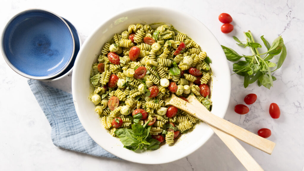 A pasta salad in a large salad bowl, with small tomatoes, basil pesto sauce, mini mozzarella balls, and chopped fresh basil. Salad serving spoons sit in the bowl.