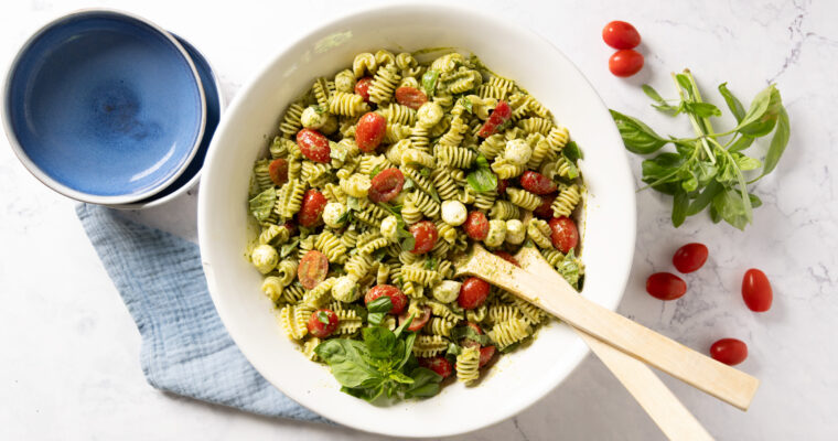 A pasta salad in a large salad bowl, with small tomatoes, basil pesto sauce, mini mozzarella balls, and chopped fresh basil. Salad serving spoons sit in the bowl.