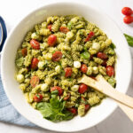 A pasta salad in a large salad bowl, with small tomatoes, basil pesto sauce, mini mozzarella balls, and chopped fresh basil. Salad serving spoons sit in the bowl.