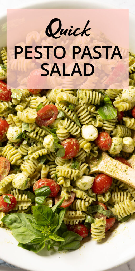 A pasta salad in a large salad bowl, with small tomatoes, basil pesto sauce, mini mozzarella balls, and chopped fresh basil. Salad serving spoons sit in the bowl.