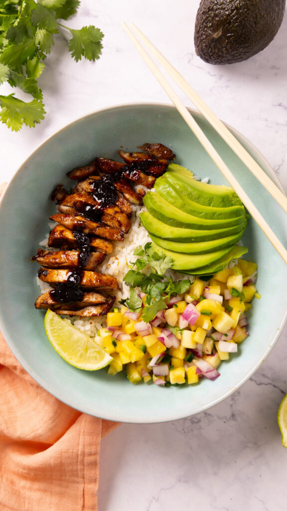 A bowl contains rice, glazed chicken, mango pineapple salsa, sliced avocado. Chopsticks sit on the edge of the bowl, surrounded by a pink napkin, limes, an avocado and a bunch of fresh cilantro.
