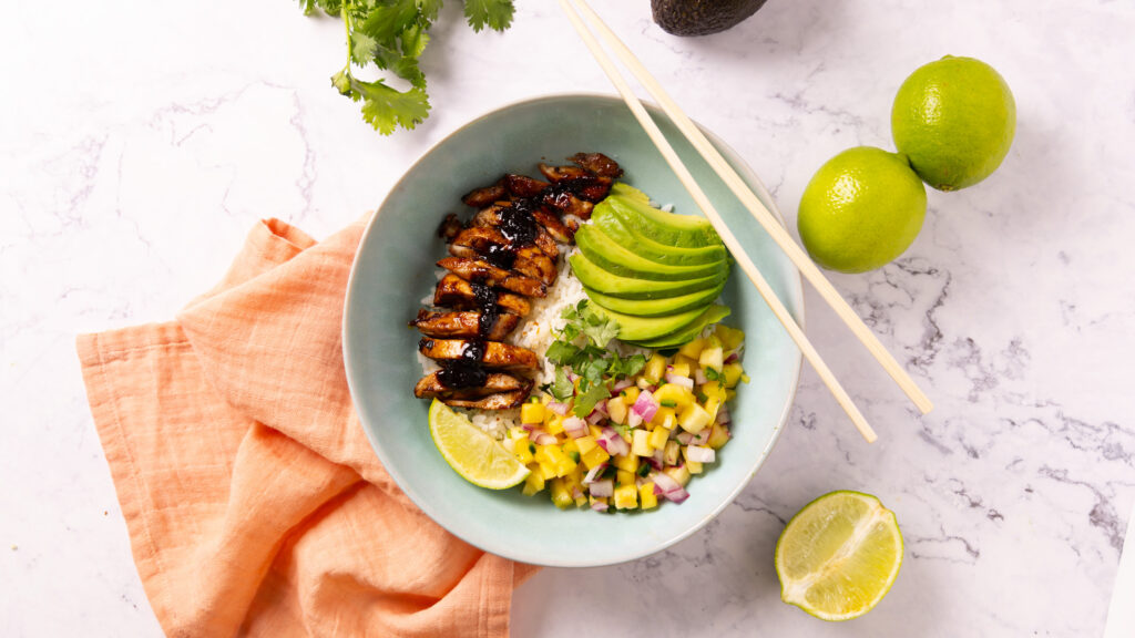 A bowl contains rice, glazed chicken, mango pineapple salsa, sliced avocado. Chopsticks sit on the edge of the bowl, surrounded by a pink napkin, limes, an avocado and a bunch of fresh cilantro.