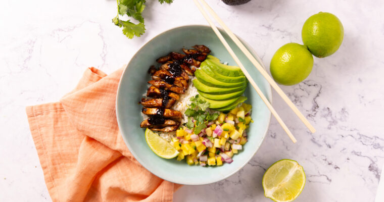 A bowl contains rice, glazed chicken, mango pineapple salsa, sliced avocado. Chopsticks sit on the edge of the bowl, surrounded by a pink napkin, limes, an avocado and a bunch of fresh cilantro.