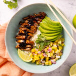 A bowl contains rice, glazed chicken, mango pineapple salsa, sliced avocado. Chopsticks sit on the edge of the bowl, surrounded by a pink napkin, limes, an avocado and a bunch of fresh cilantro.