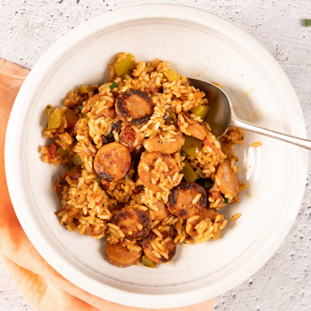 A bowl holds a chorizo sausage and rice dish. To the side, A large skillet holds a chorizo sausage and rice dish. A large wooden spoon is in the skillet.