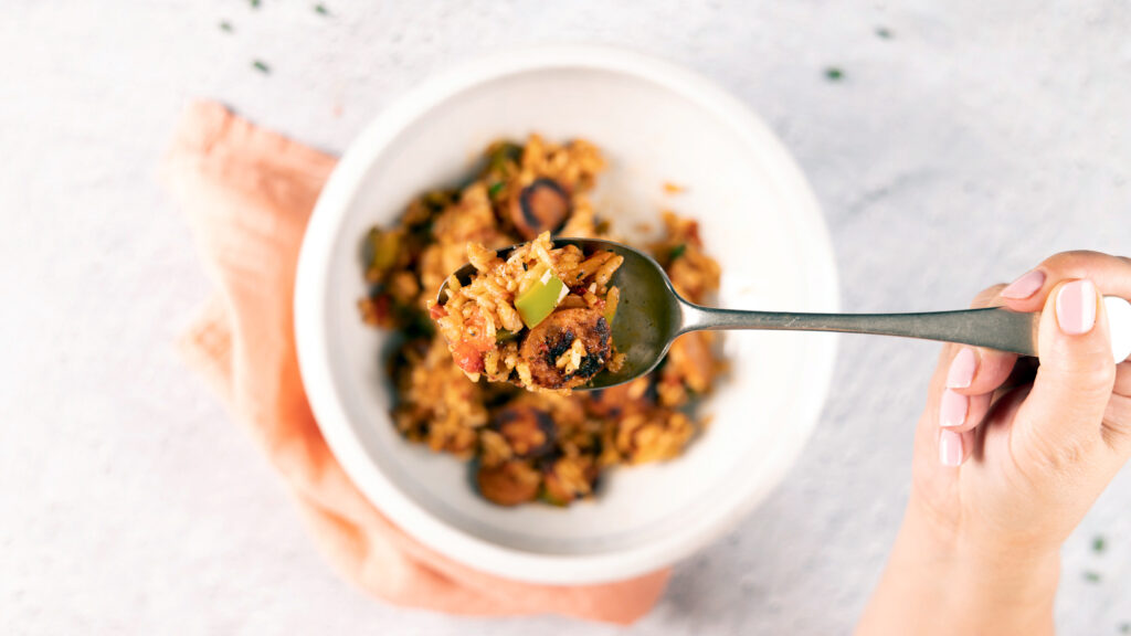 A spoon holds a bite of chorizo sausage and rice dish, above a bowl. To the side, A large skillet holds a chorizo sausage and rice dish. A large wooden spoon is in the skillet.