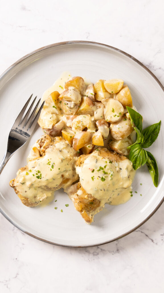 A plate with two chicken thighs and a pile of baby potatoes, covered in a creamy Dijon mustard sauce, and a fork.