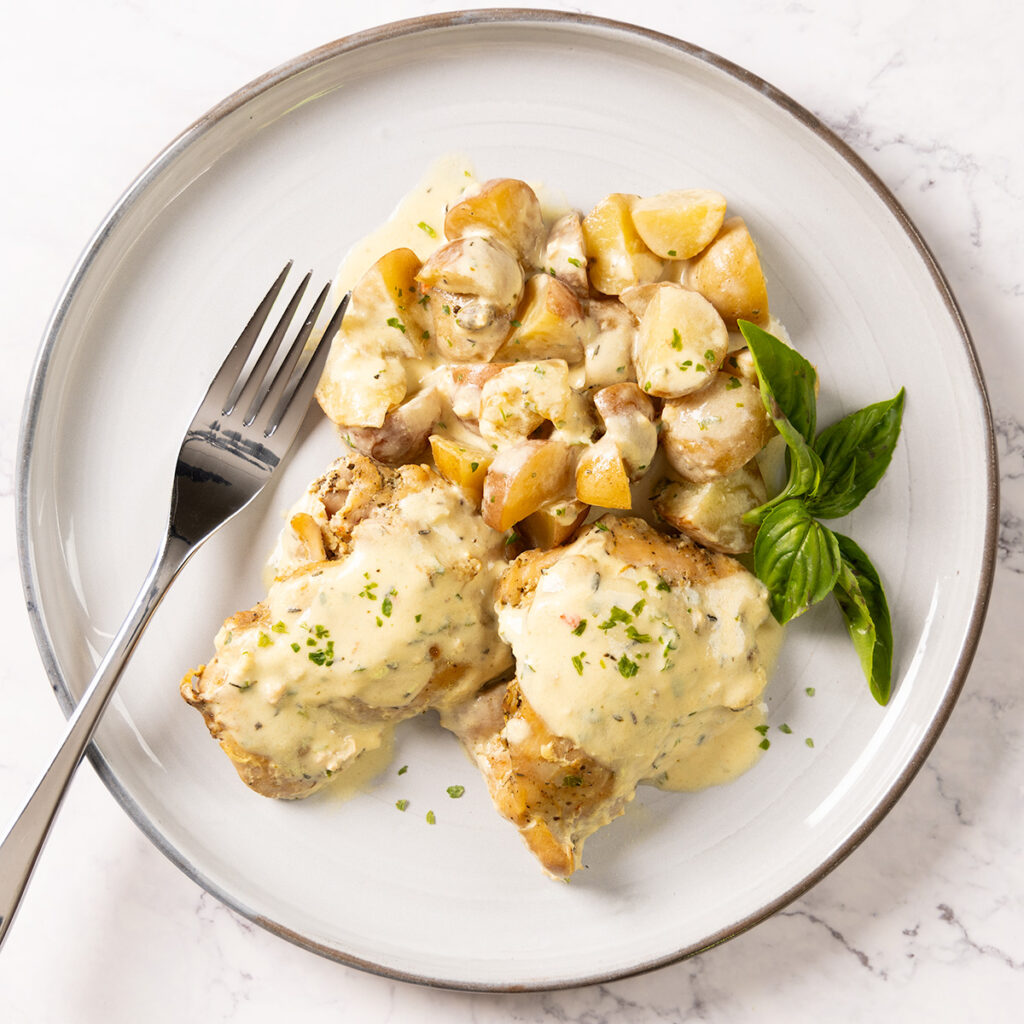 A plate with two chicken thighs and a pile of baby potatoes, covered in a creamy Dijon mustard sauce, and a fork.