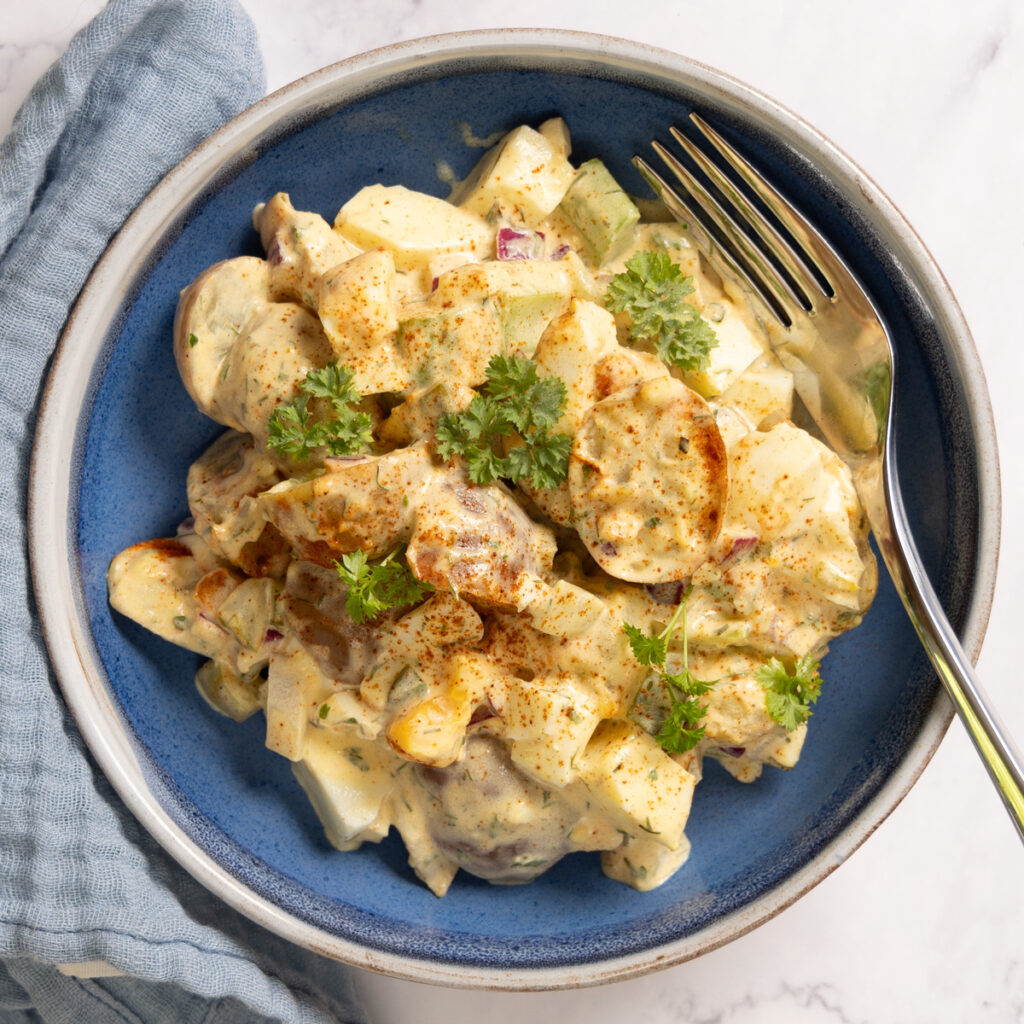 A bowl contains potato salad, and a fork. A blue napkin is next to the bowl. 