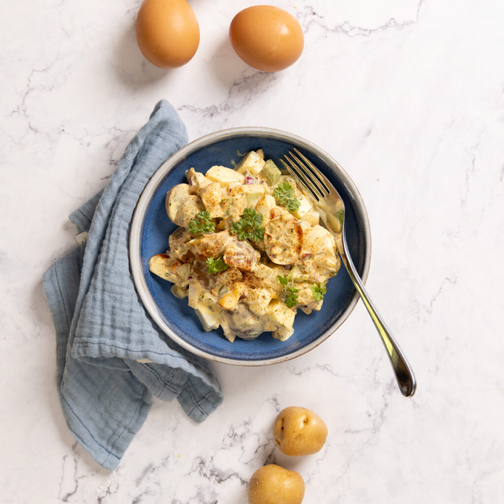 A bowl contains potato salad, and a fork. A blue napkin is next to the bowl. 2 boiled eggs and 2 potatoes are next to the bowl.