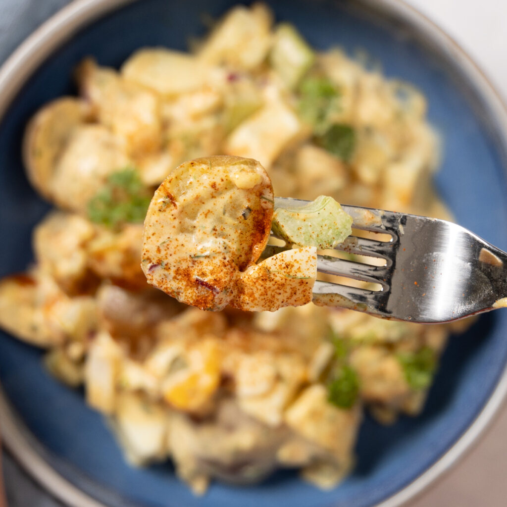 A bowl contains potato salad. A hand lifts a forkful of potato salad up to the camera. 