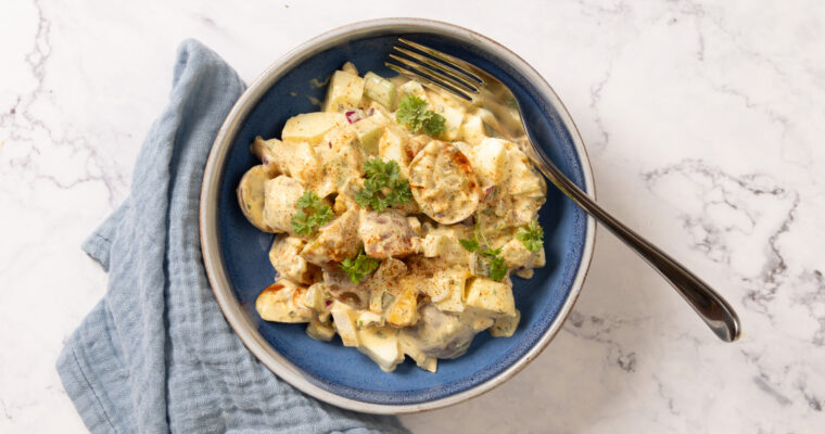 A bowl contains potato salad, and a fork. A blue napkin is next to the bowl.