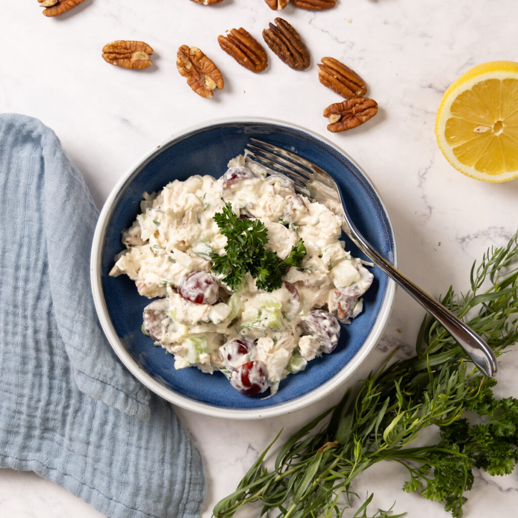 A small bowl contains chicken salad. In the salad is creamy dressing, red grapes, chunks of chicken, bits of celery, onion, and herbs.  Next to the bowl are pecans, bunches of herbs, a cut lemon, and red grapes. A blue napkin is next the bowl.