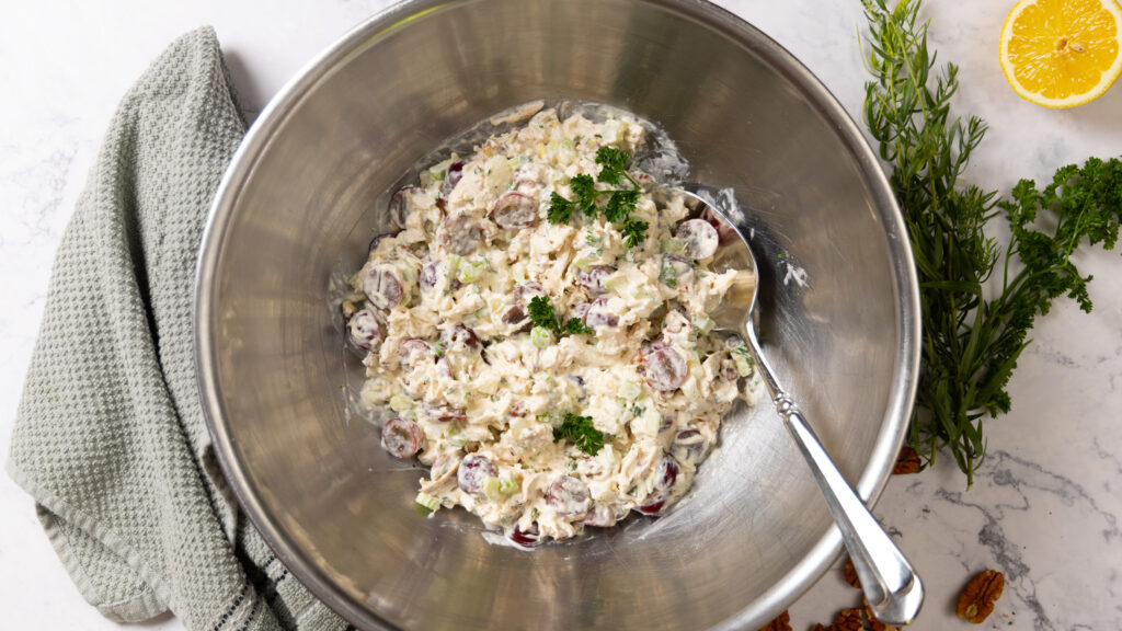 A stainless steel mixing bowl contains a freshly mixed chicken salad containing chunks of chicken, red grapes, celery, onions, pecans, and herbs. A large mixing spoon is also in the bowl.