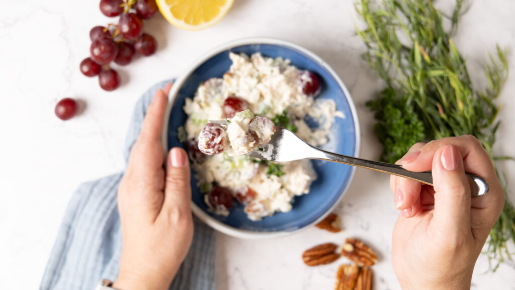 A small bowl contains chicken salad. A hand holding a fork is holding up a bite of salad; in the salad is creamy dressing, red grapes, chunks of chicken, bits of celery, onion, and herbs. Next to the bowl are pecans, bunches of herbs, a cut lemon, and red grapes. A blue napkin is next the bowl.