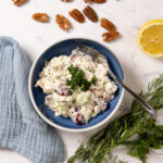 A small bowl contains chicken salad. In the salad is creamy dressing, red grapes, chunks of chicken, bits of celery, onion, and herbs. Next to the bowl are pecans, bunches of herbs, a cut lemon, and red grapes. A blue napkin is next the bowl.