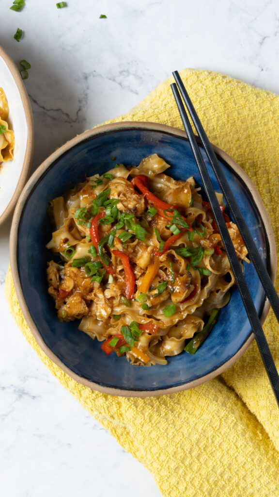 A bowl of Chili Garlic Chicken Noodle stir-fry, with black chopsticks, a yellow towel, and green onions next to the bowl.