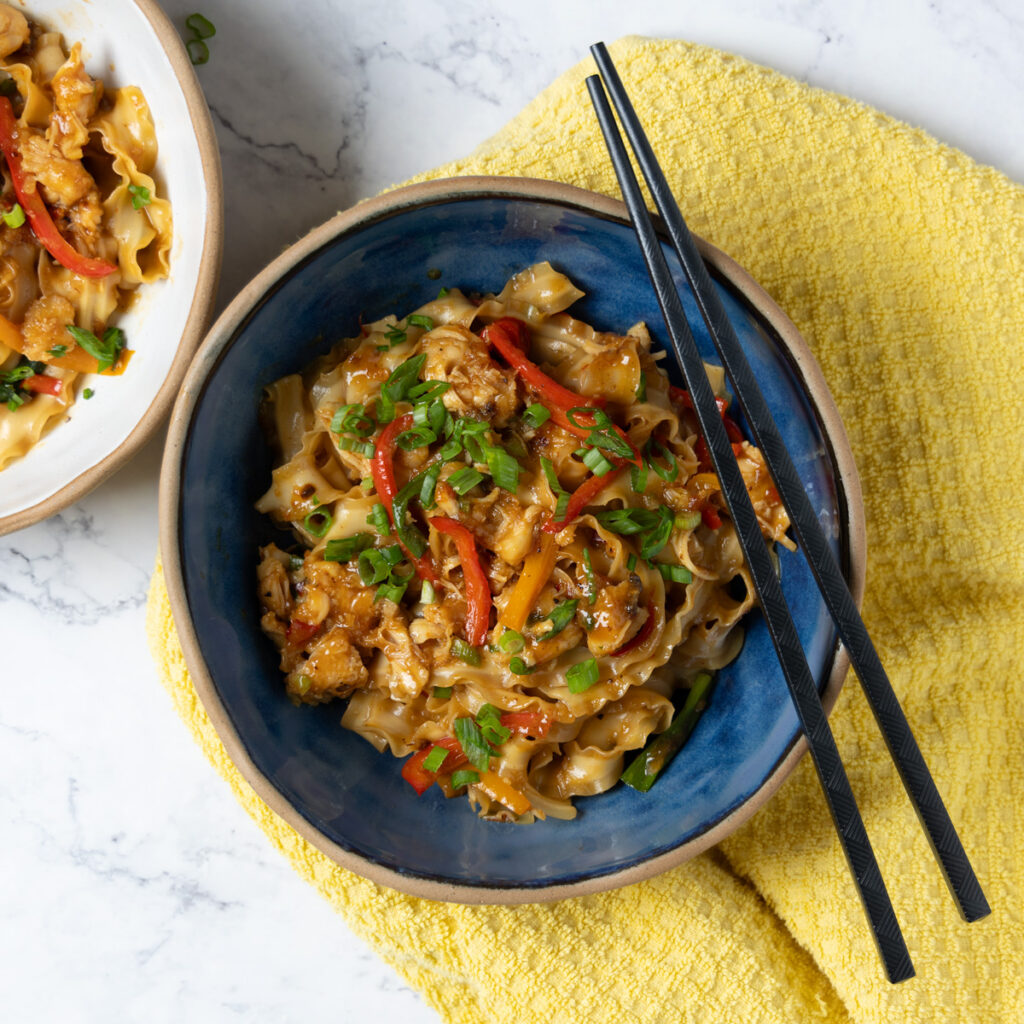 A bowl of Chili Garlic Chicken Noodle stir-fry, with black chopsticks, a yellow towel, and green onions next to the bowl.