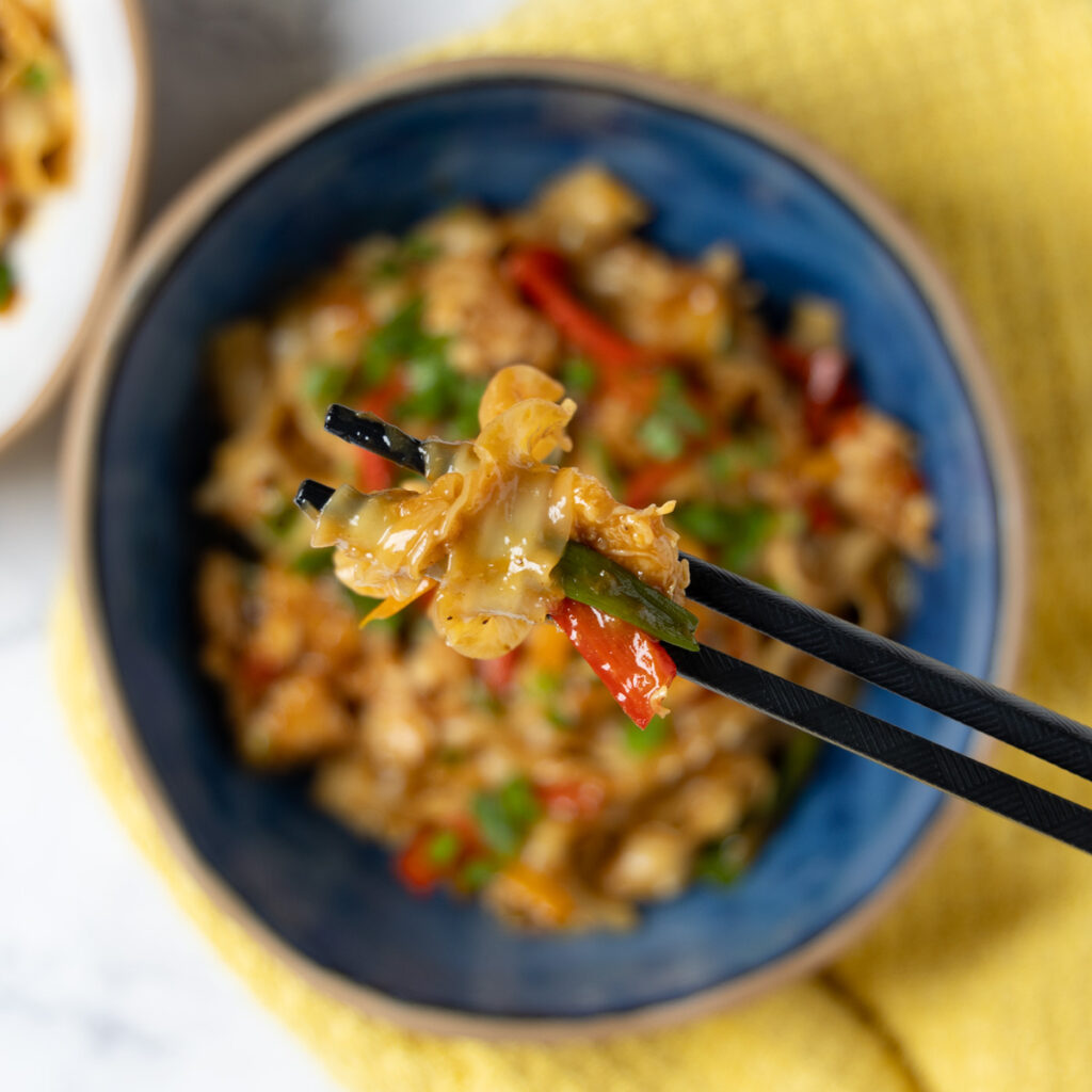 A bowl of Chili Garlic Chicken Noodle stir-fry, with black chopsticks, a yellow towel, and green onions next to the bowl.