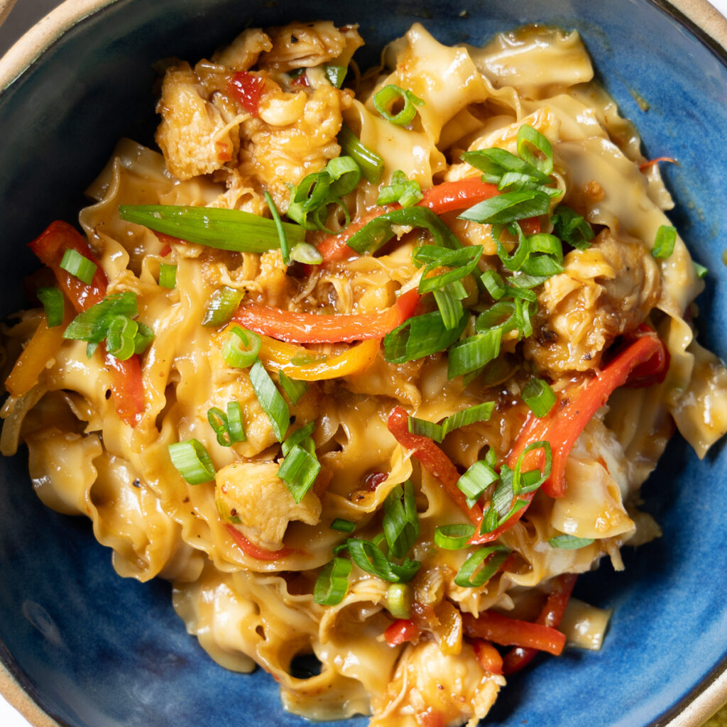 A bowl of Chili Garlic Chicken Noodle stir-fry, with black chopsticks, a yellow towel, and green onions next to the bowl.