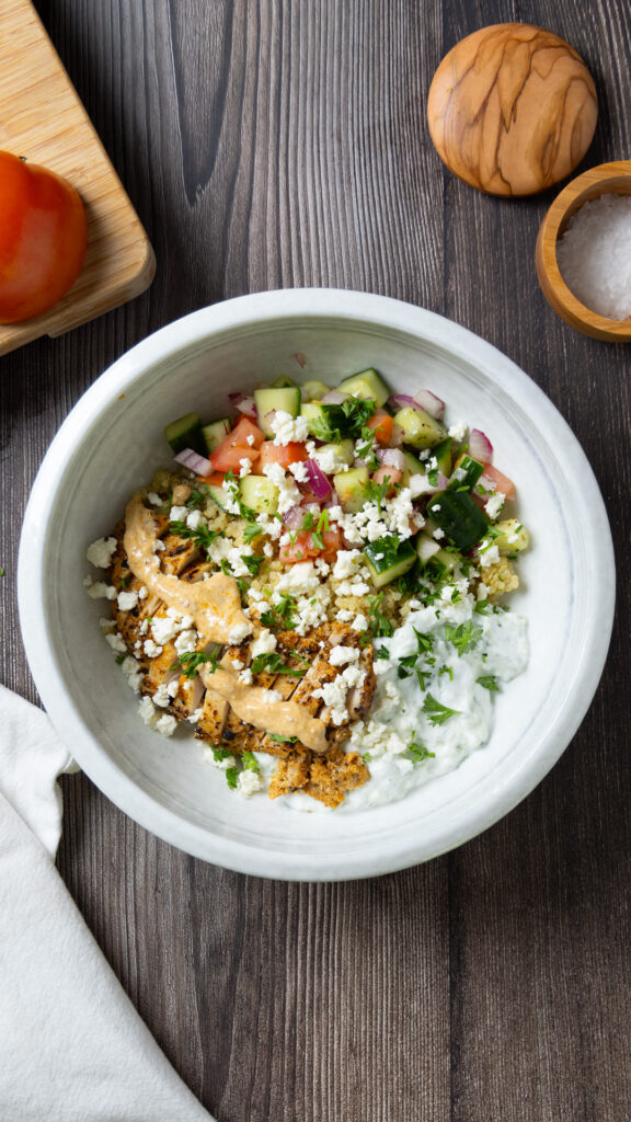 A Greek Chicken Power Bowl, on a wooden table, on the table is a small cutting board with a tomato, a wooden bowl of salt, a white napkin, a fork, a wooden spoon, some lemon wedges, a red onion, and some parsley.