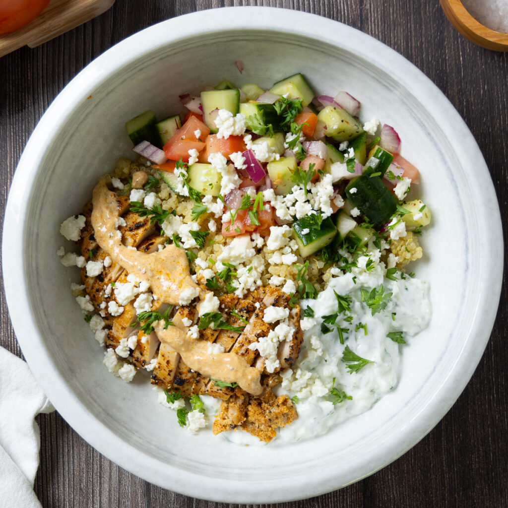 A Greek Chicken Power Bowl, on a wooden table, on the table is a small cutting board with a tomato, a wooden bowl of salt, a white napkin, a fork, a wooden spoon, some lemon wedges, a red onion, and some parsley.