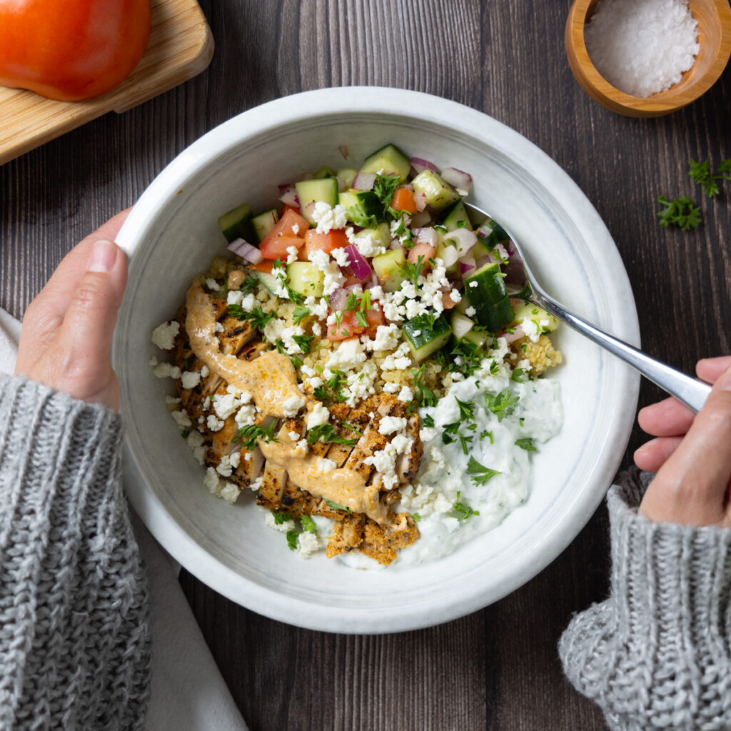 A Greek Chicken Power Bowl, on a wooden table, on the table is a small cutting board with a tomato, a wooden bowl of salt, a white napkin, a fork, a wooden spoon, some lemon wedges, a red onion, and some parsley. The bowl is held by hands.