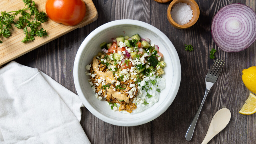 A Greek Chicken Power Bowl, on a wooden table, on the table is a small cutting board with a tomato, a wooden bowl of salt, a white napkin, a fork, a wooden spoon, some lemon wedges, a red onion, and some parsley.