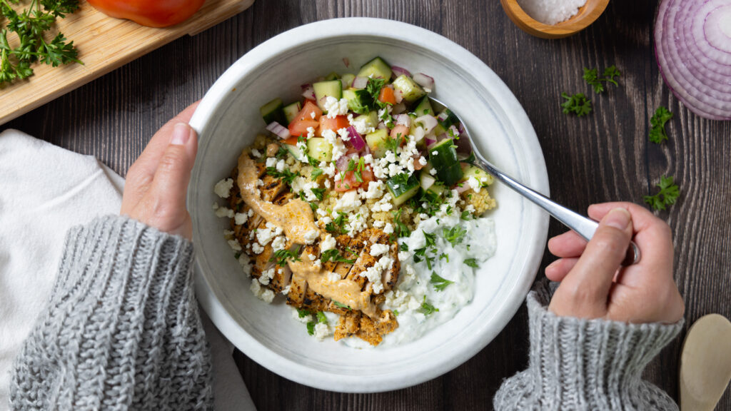 A Greek Chicken Power Bowl, on a wooden table, on the table is a small cutting board with a tomato, a wooden bowl of salt, a white napkin, a fork, a wooden spoon, some lemon wedges, a red onion, and some parsley.Hands are holding the bowl.