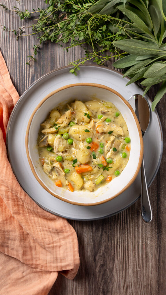 A bowl sits on a plate, inside the bowl is a dish called Chicken Pot Pie Gnocchi; carrots, celery and mushrooms in a creamy sauce with gnocchi. Some herbs are near the plate. A napkin is nearby.