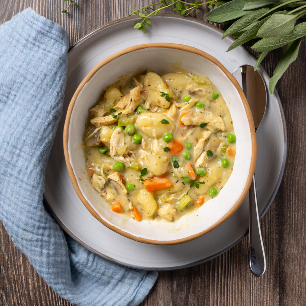 A bowl sits on a plate, inside the bowl is a dish with Chicken and Gnocchi; carrots, celery and mushrooms in a creamy sauce with gnocchi. Some herbs are near the plate. A napkin is nearby.